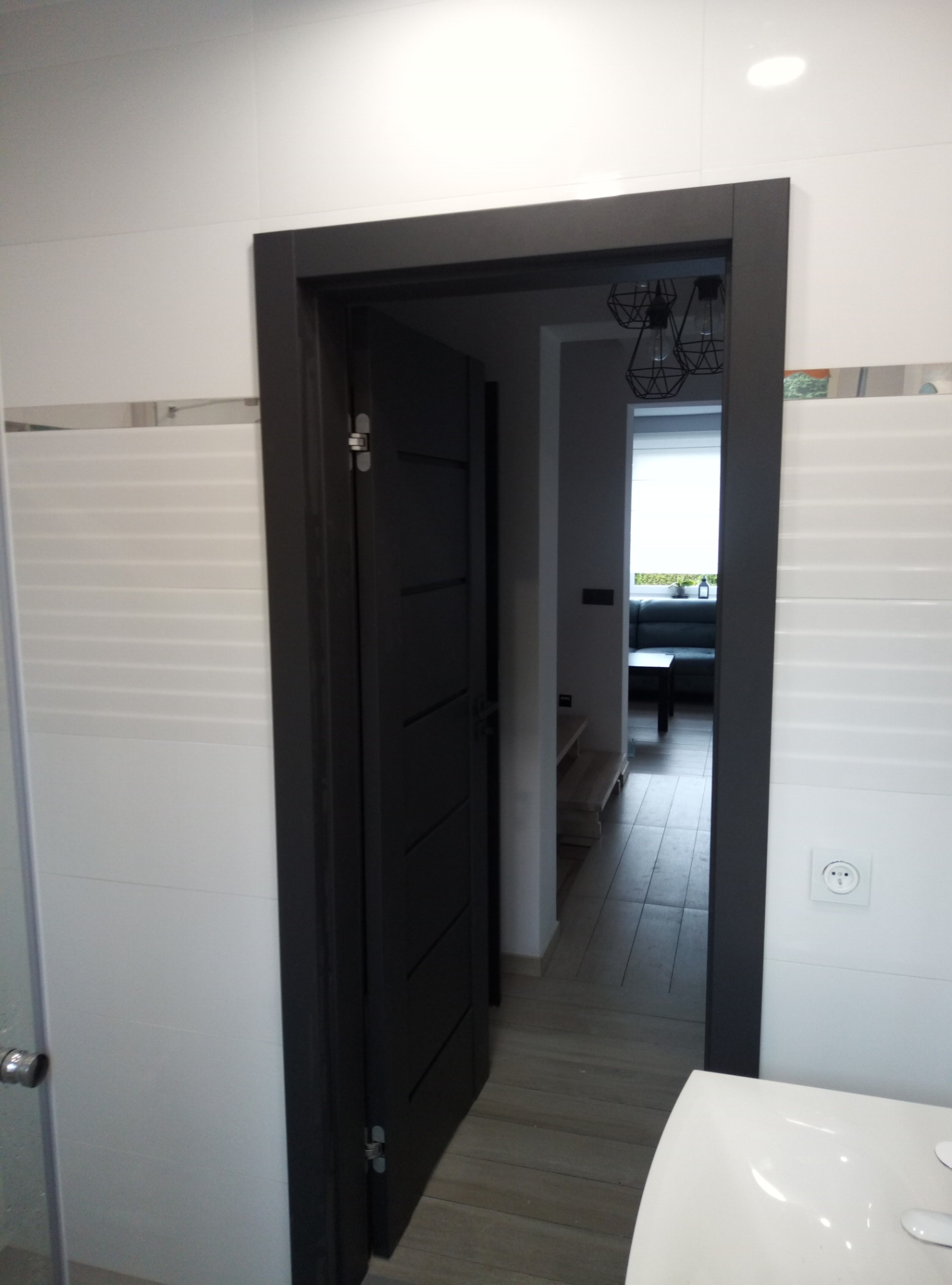 Otwarta, czarna, wewnętrzna stolarka drzwiowa z widokiem na salon z szarą sofą, widziana z łazienki z białymi płytkami i umywalką.