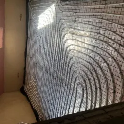 Białe rurki układane spiralnie na czarnej siatce, prawdopodobnie system ogrzewania podłogowego w trakcie instalacji w pomieszczeniu z surowymi ścianami.
