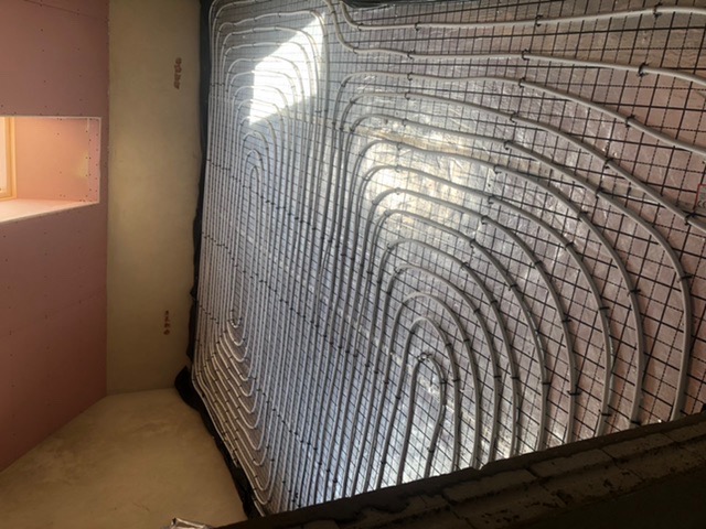 Białe rurki układane spiralnie na czarnej siatce, prawdopodobnie system ogrzewania podłogowego w trakcie instalacji w pomieszczeniu z surowymi ścianami.