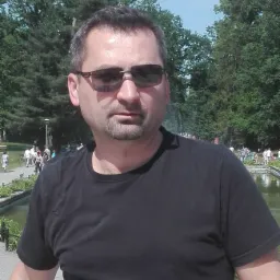 Mężczyzna w czarnej koszulce i okularach przeciwsłonecznych, stojący w parku na tle fontanny i spacerujących ludzi.