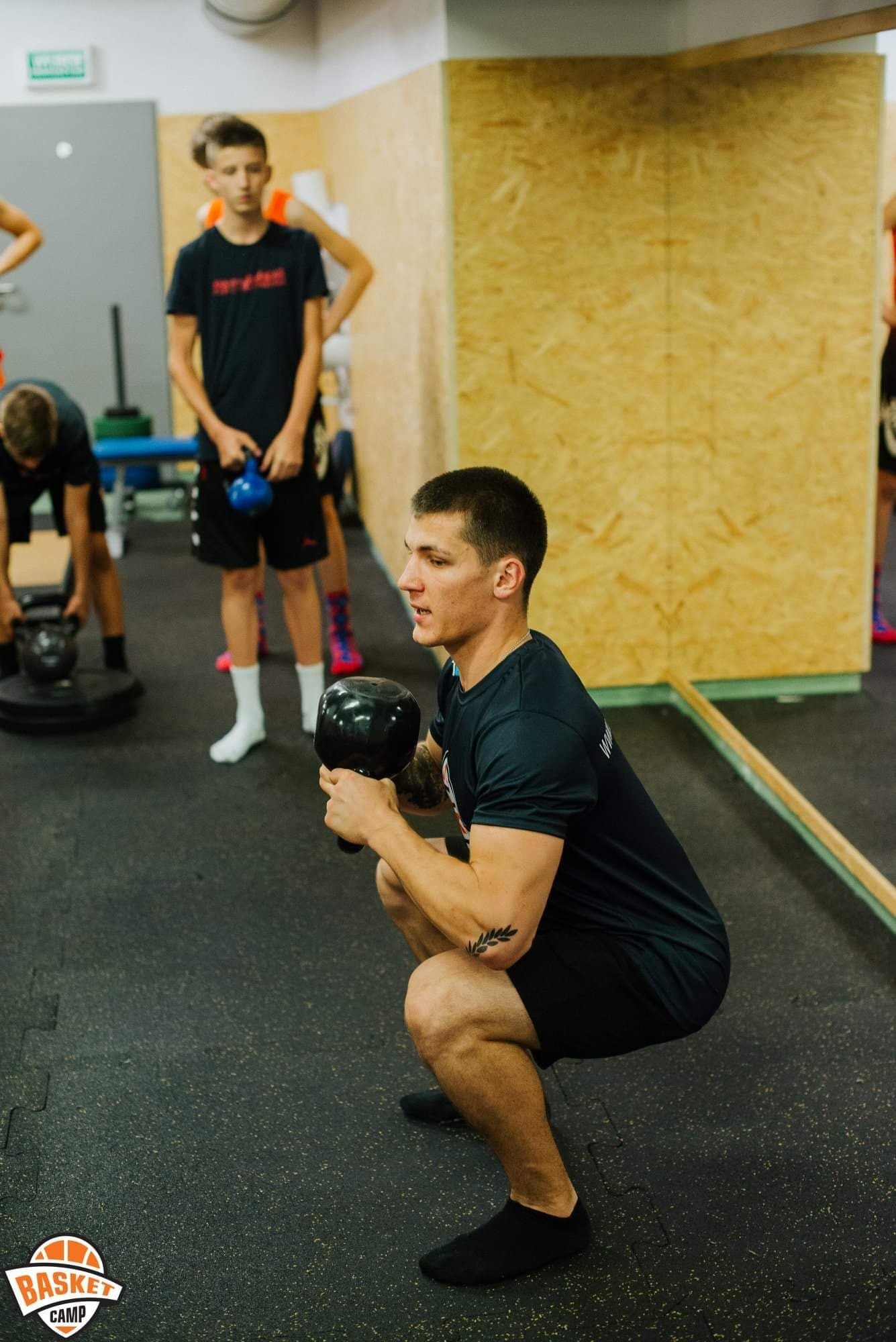 Mężczyzna w czarnym stroju sportowym wykonuje przysiad z kettlebellem w siłowni; w tle młodzież z obciążeniami.
