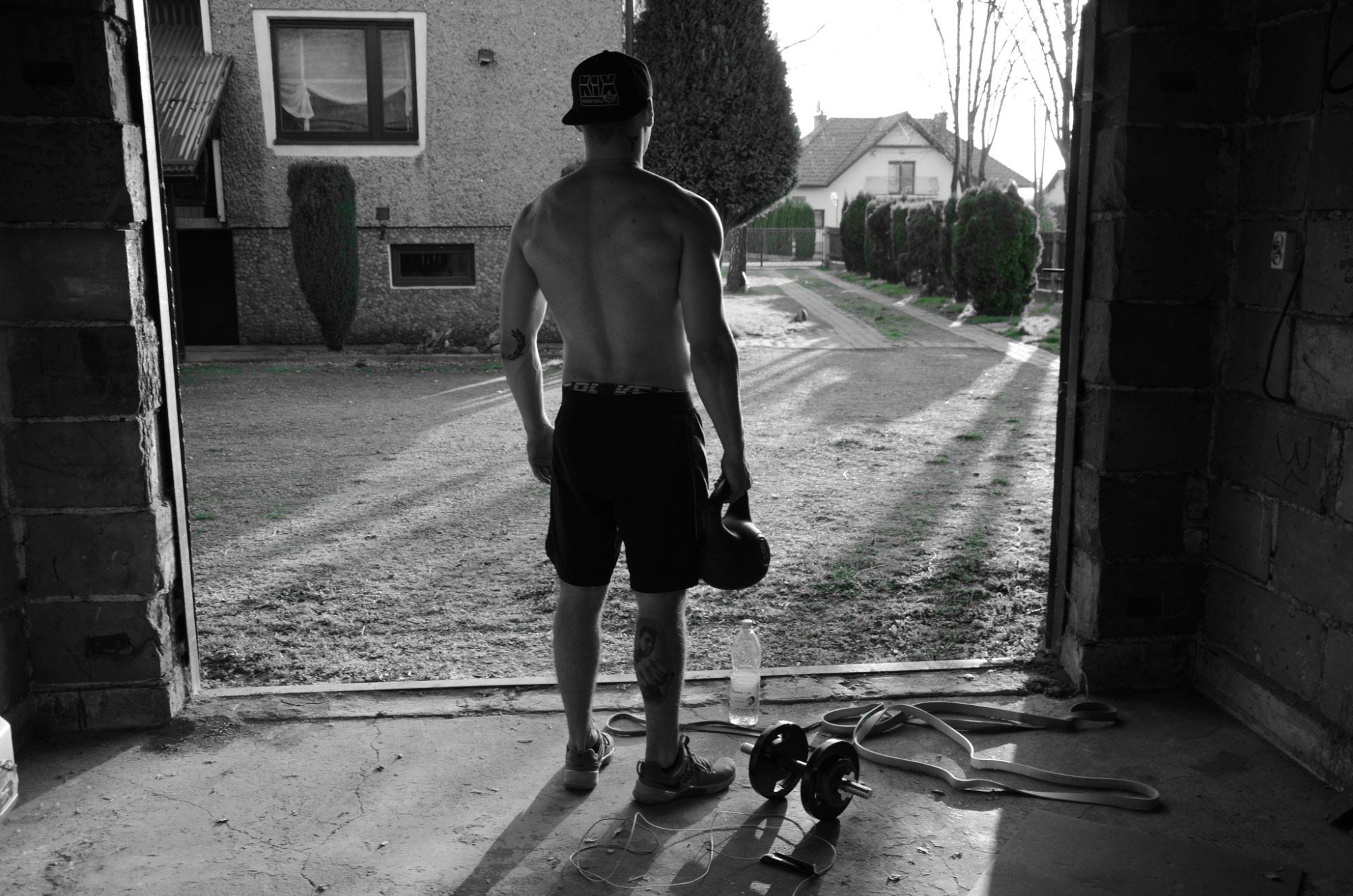 Mężczyzna tyłem, z kettlebell w dłoni, stoi w wejściu do garażu; na podłodze leżą hantle, guma do ćwiczeń i butelka wody. Widok na ogród i domy w tle, w sepii.