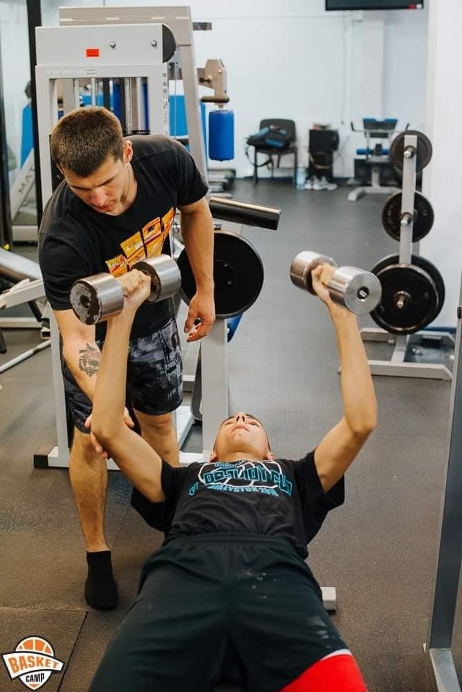 Młody mężczyzna leżący na ławeczce, podnoszący hantle pod nadzorem drugiego mężczyzny w siłowni, z widocznym tatuażem na ramieniu nadzorującego.