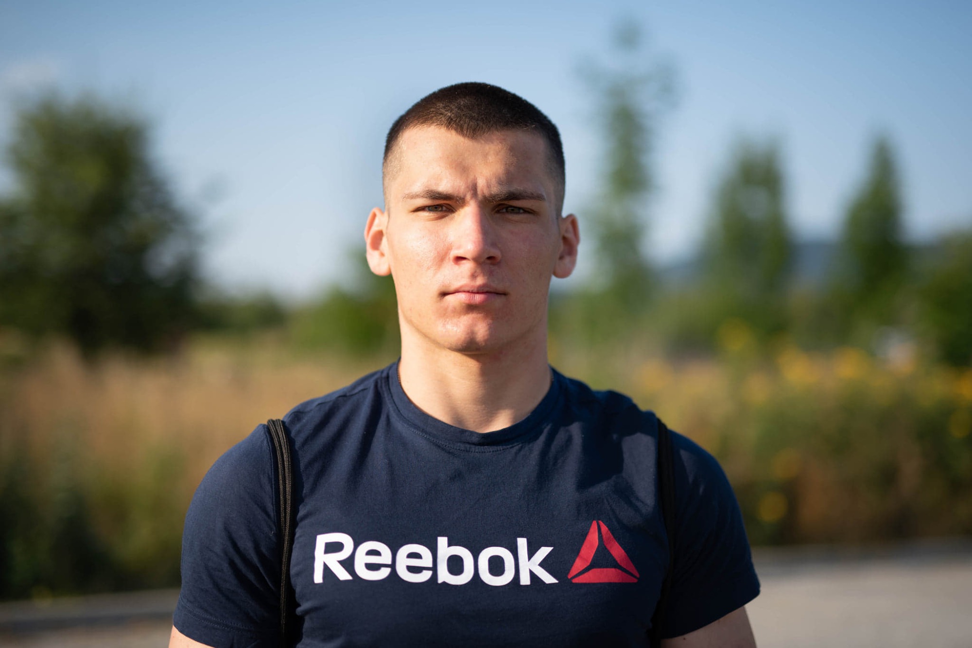Portret młodego mężczyzny w granatowej koszulce Reebok, z krótkimi włosami, na rozmytym tle zieleni i błękitnego nieba.