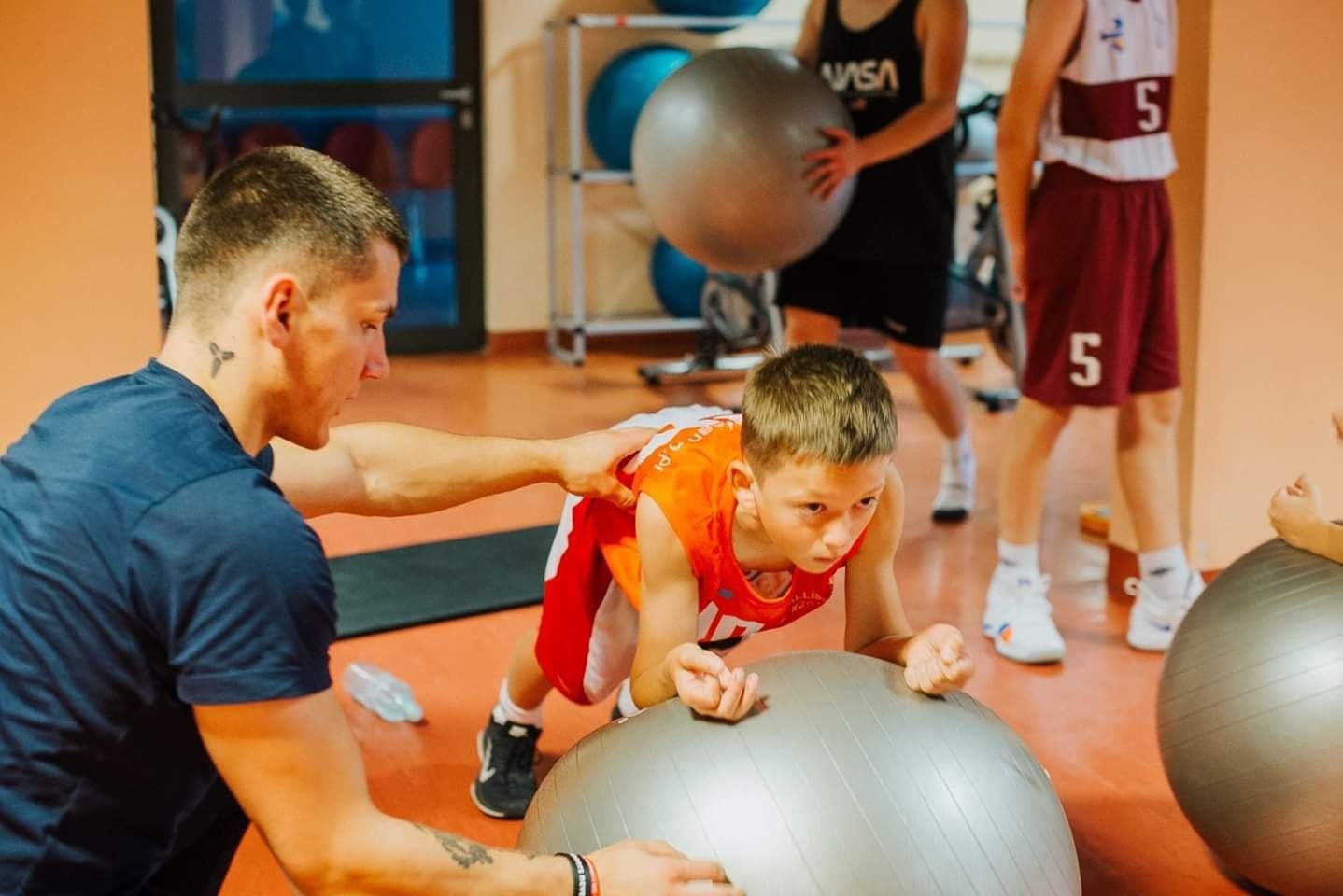 Chłopiec w stroju sportowym wykonuje ćwiczenie na piłce rehabilitacyjnej pod nadzorem mężczyzny z tatuażem na szyi w siłowni.