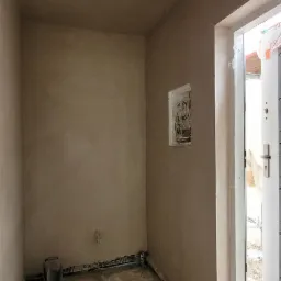 Wnętrze pomieszczenia po nałożeniu tynku, widoczne przewody elektryczne wystające ze ściany i zwój kabla na podłodze, otwarte drzwi.