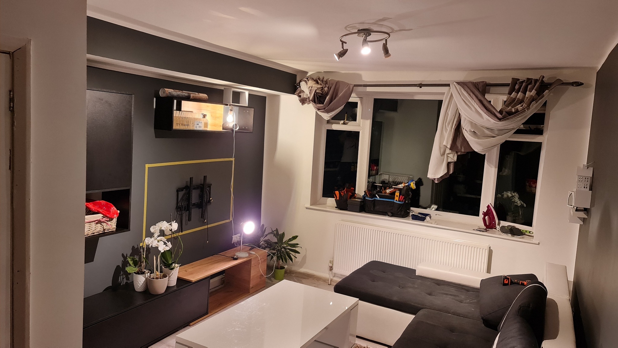 Salon w trakcie prac wykończeniowych: zamontowany uchwyt TV na ciemnej ścianie z żółtą taśmą malarską, narzędzia na parapecie, widoczne elementy umeblowania i oświetlenia.