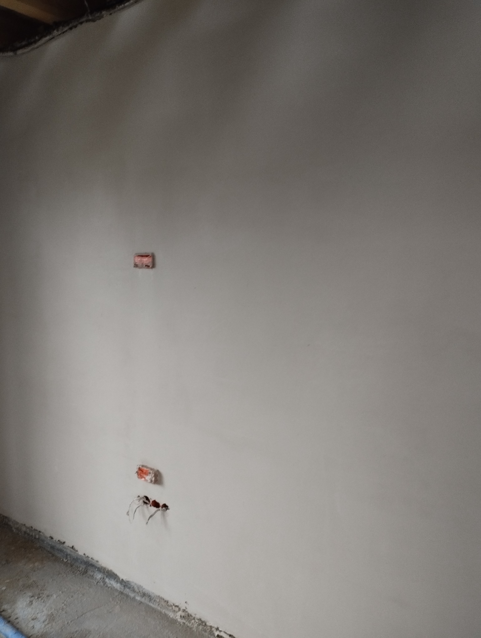 Świeżo nałożony, gładki tynk maszynowy na ścianie, z widocznymi puszkami instalacyjnymi i wystającymi przewodami elektrycznymi. W tle widoczne fragmenty surowego sufitu.