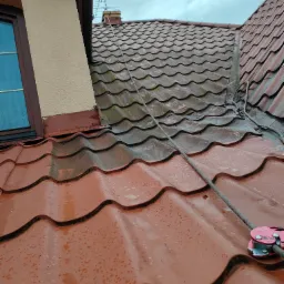 Czerwona dachówka karpiówka, częściowo mokra po deszczu, z liną zabezpieczającą i przyrządem zaciskowym, widoczny fragment okna dachowego i ściany budynku.