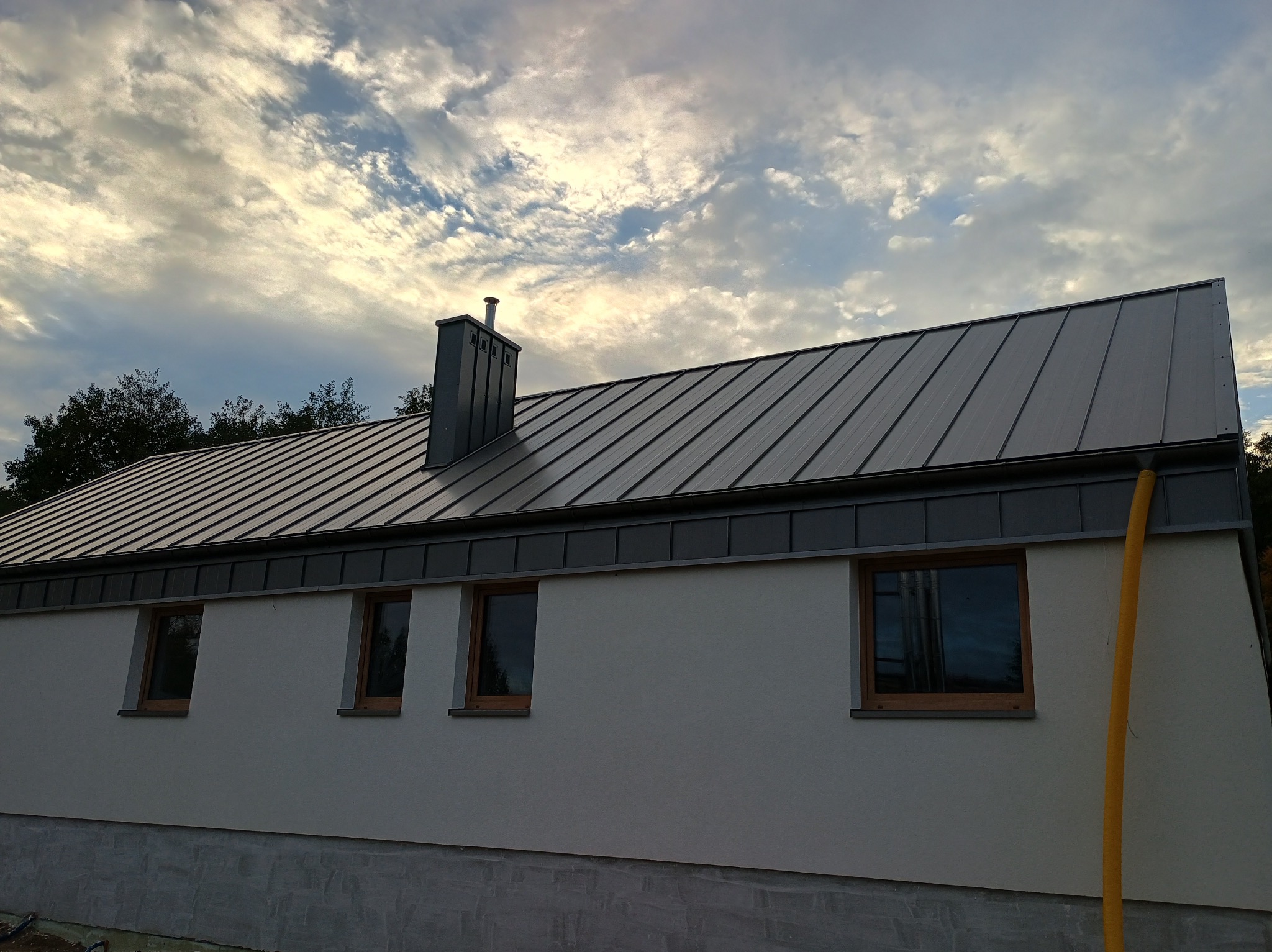 Nowoczesny dom z szarą, metalową dachówką trapezową, widok na elewację z czterema oknami i kominem na tle pochmurnego nieba.