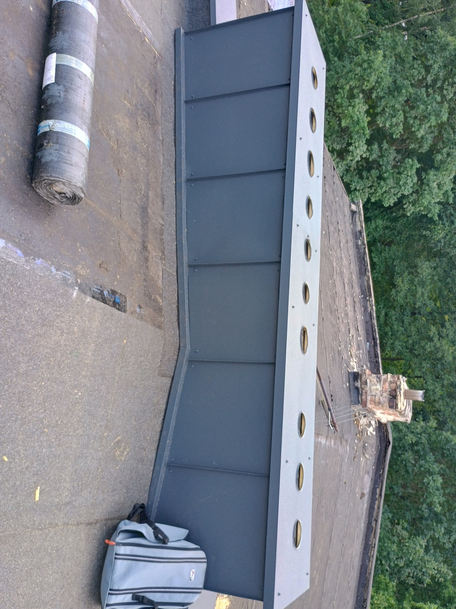 Widok z góry na płaski dach z nową, szarą obróbką blacharską z okrągłymi otworami wentylacyjnymi, zwój papy, torba narzędziowa i komin w tle.