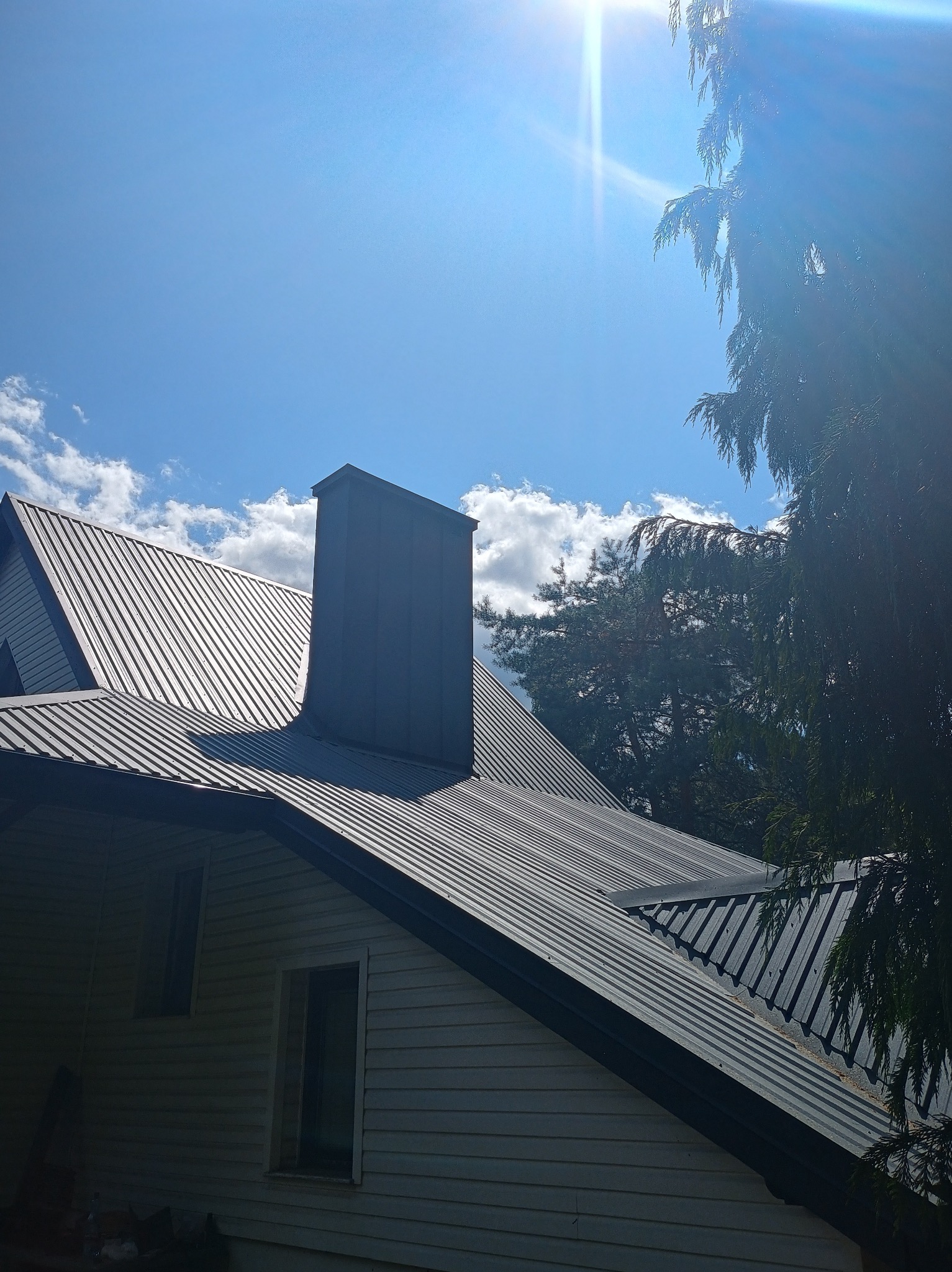 Dom z szarym dachem z blachy trapezowej i kominem, widok z dołu w słoneczny dzień z drzewami w tle.