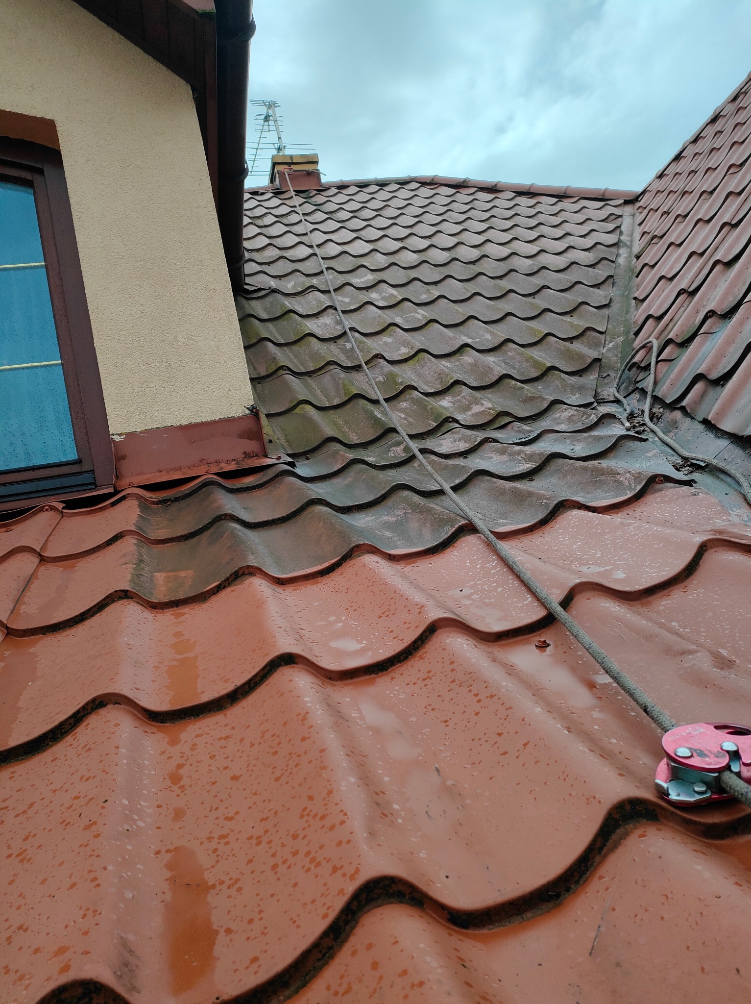 Czerwona dachówka karpiówka, częściowo mokra po deszczu, z liną zabezpieczającą i przyrządem zaciskowym, widoczny fragment okna dachowego i ściany budynku.
