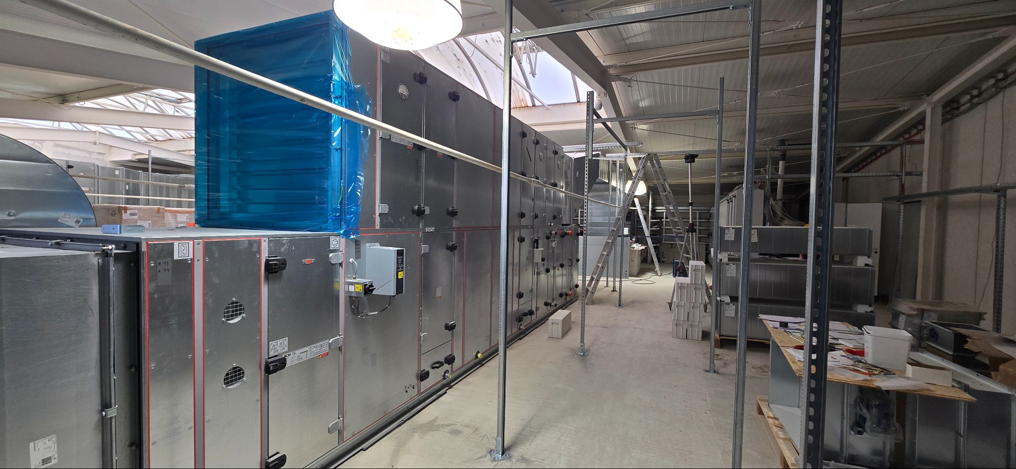 Długa ściana z modułów wentylacyjnych w hali, częściowo zabezpieczona folią, widoczne drabiny i elementy konstrukcyjne sufitu.