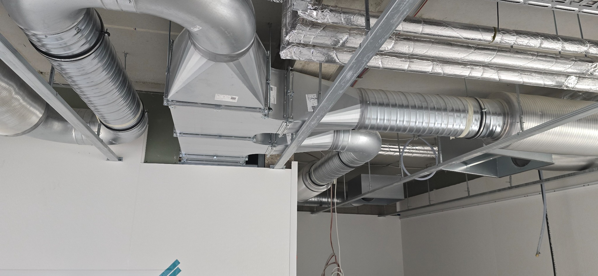 Metalowe rury wentylacyjne pod sufitem, widoczne elementy izolacji termicznej i mocowania.
