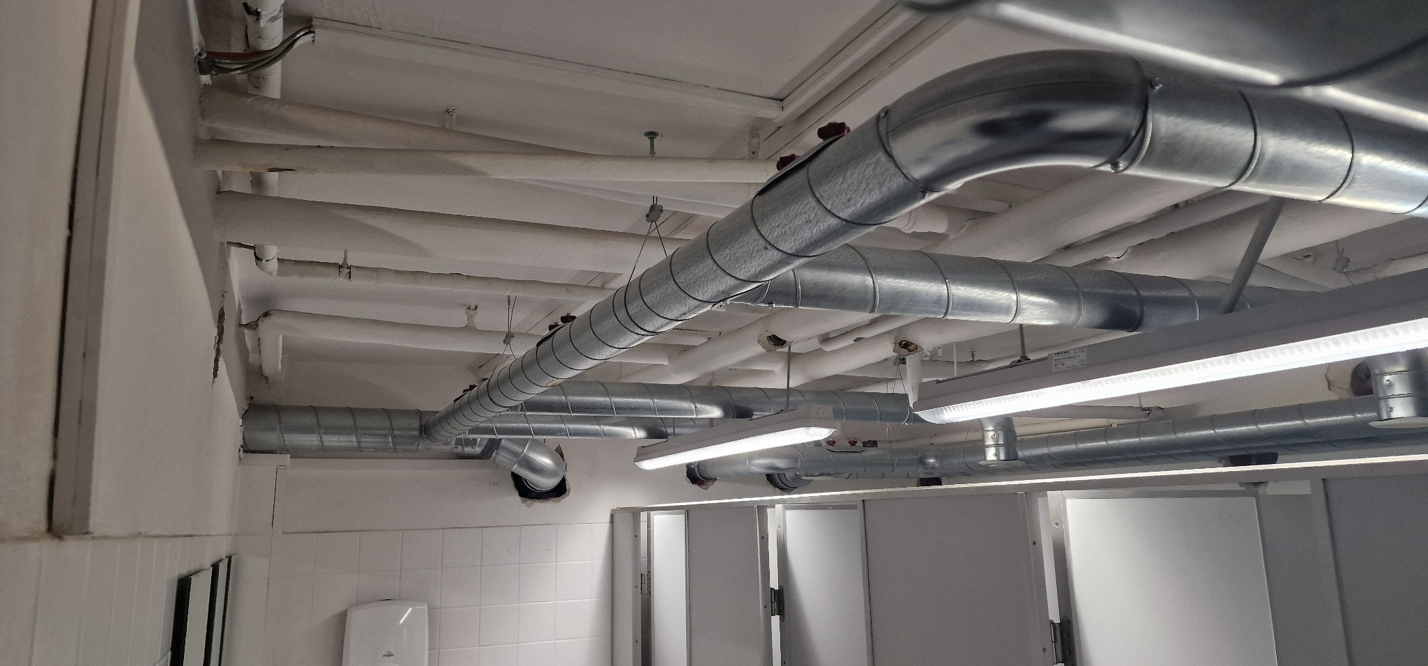 Metalowe rury wentylacyjne pod sufitem w pomieszczeniu z oświetleniem LED i białymi ścianami.