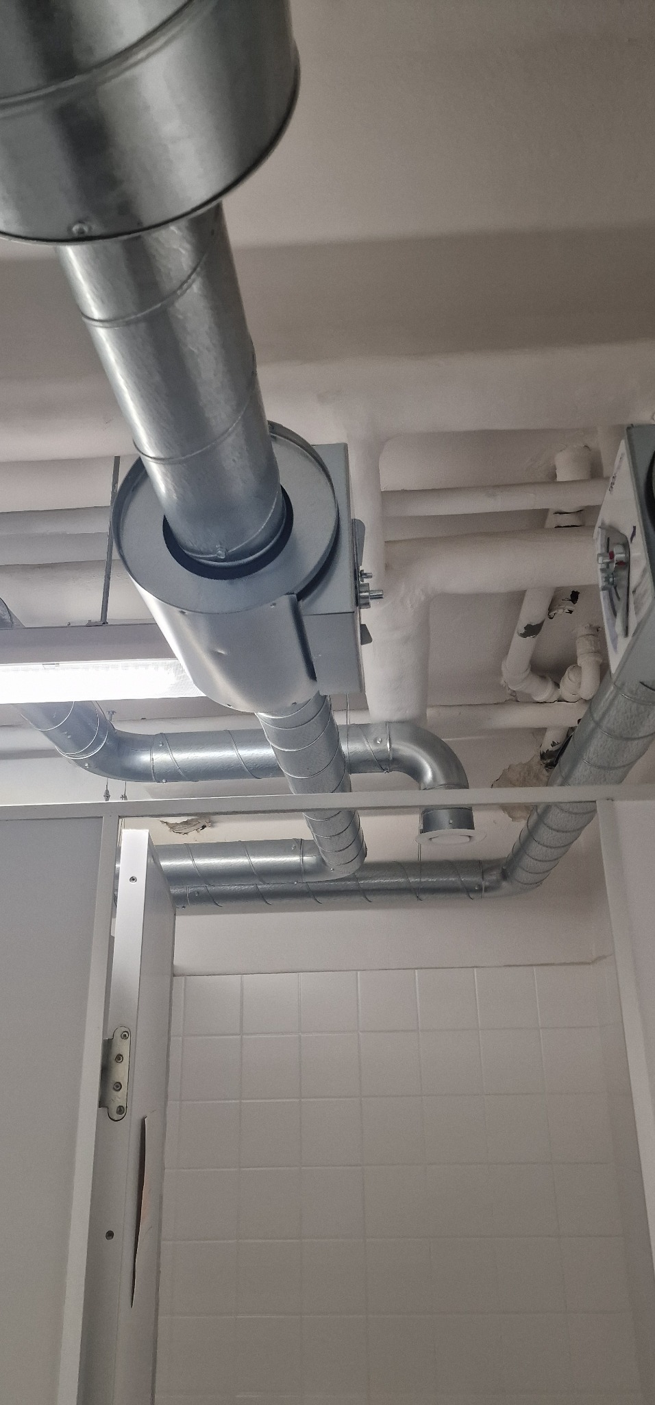 Widok z dołu na zainstalowany system wentylacyjny z rurami i elementami mocującymi w pomieszczeniu w Wiehl, widoczna biała ściana z płytkami i fragment drzwi.