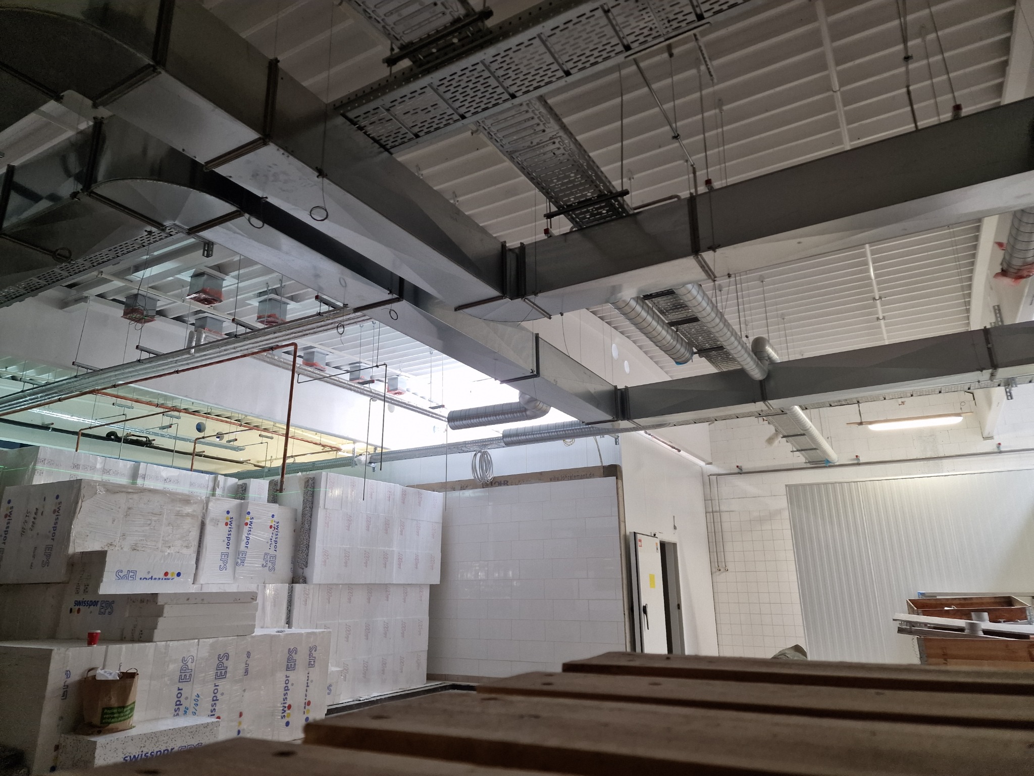 Widok na sufit hali przemysłowej z zainstalowaną, rozbudowaną siecią kanałów wentylacyjnych, rur i korytek kablowych, poniżej składowane paczki styropianu EPS.