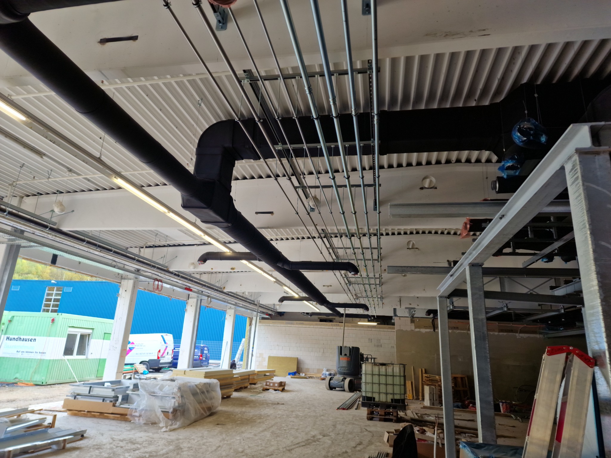 Wnętrze hali w trakcie instalacji systemu wentylacyjnego: widoczne czarne rury wentylacyjne pod sufitem, metalowe rurki instalacyjne, materiały budowlane składowane na podłodze oraz kontener...