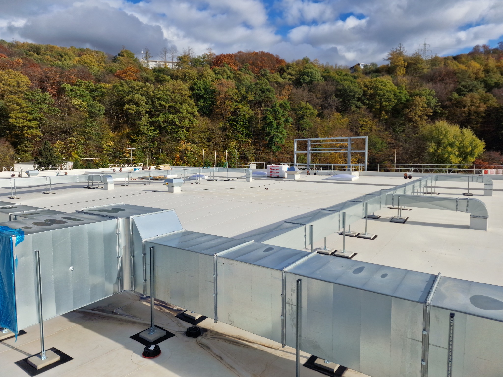 System kanałów wentylacyjnych z blachy ocynkowanej zamontowany na dachu budynku przemysłowego, widok z góry, w tle zalesione wzgórze w jesiennej kolorystyce.