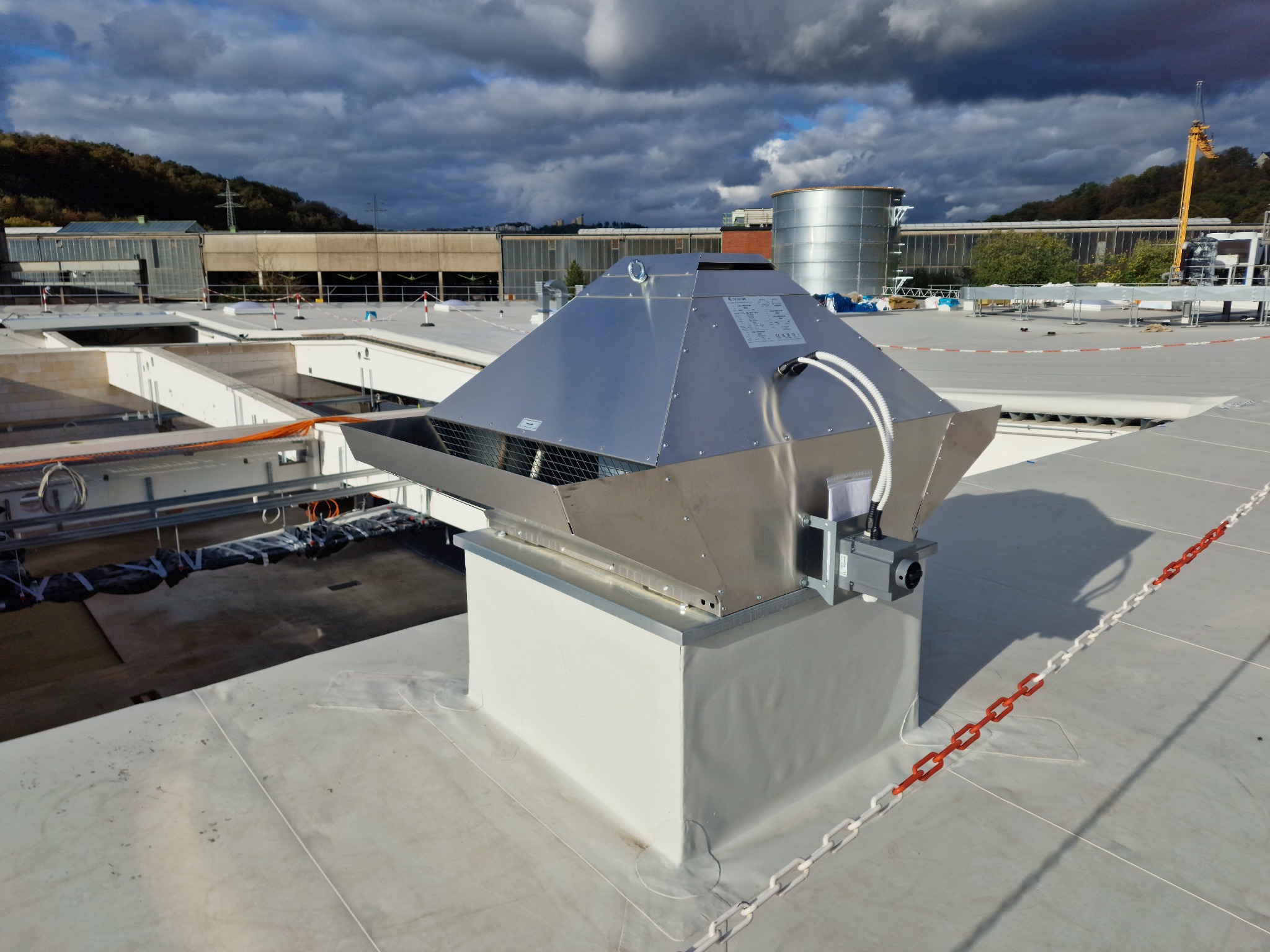 Pirymidalny, metalowy wywietrznik dachowy z kratką ochronną, zamontowany na białej podstawie, widoczny na tle przemysłowego krajobrazu i pochmurnego nieba.