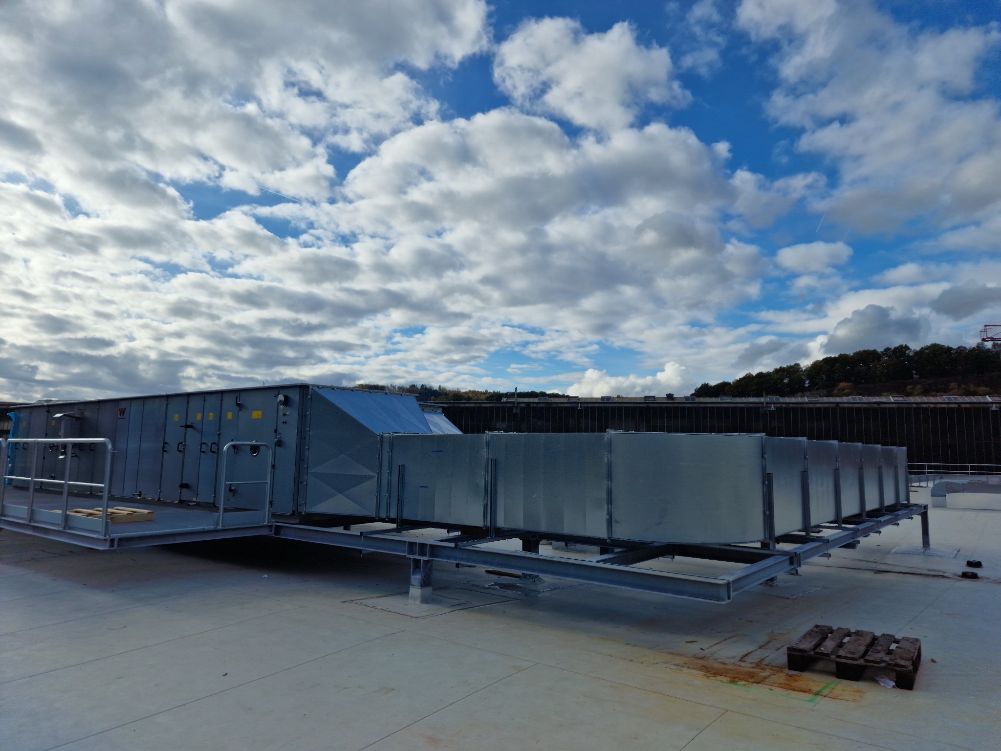 Centrala wentylacyjna na dachu budynku, widok z dołu, błękitne niebo z obłokami w tle.