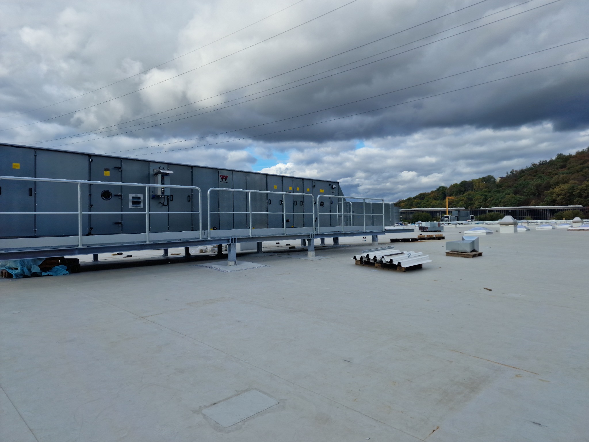 Duża centrala wentylacyjna z metalowymi barierkami na płaskim dachu budynku przemysłowego w pochmurny dzień.