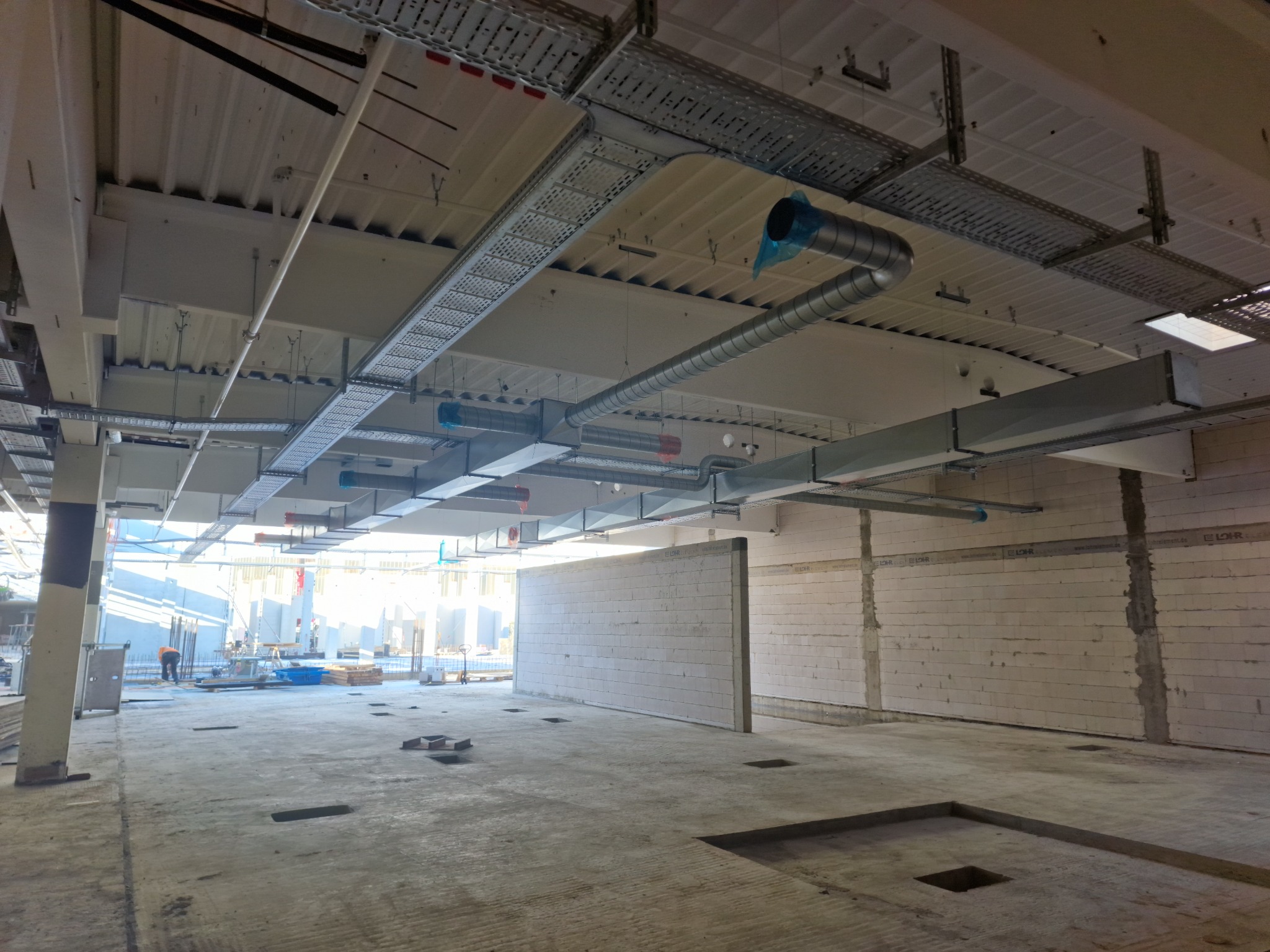 Wnętrze hali w trakcie budowy z widocznymi kanałami wentylacyjnymi pod sufitem i instalacjami elektrycznymi w korytach kablowych.