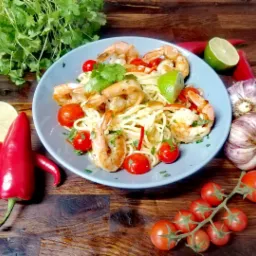 Aromatyczne danie z krewetek, makaronu spaghetti, pomidorków cherry, kolendry i limonki, podane na błękitnym talerzu, z dodatkiem papryczek chili i czosnku na drewnianym tle.