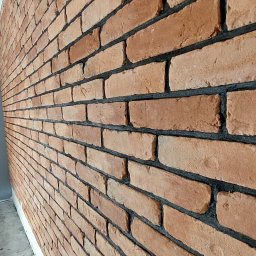 RED DIAMOND Remonty i wykończenia - Ściana wyłożona cegłą klinkierową w odcieniach czerwieni i brązu, z ciemną fugą, widoczna z perspektywy bocznej, z fragmentem szarej podłogi i białej listwy przypodłogowej.