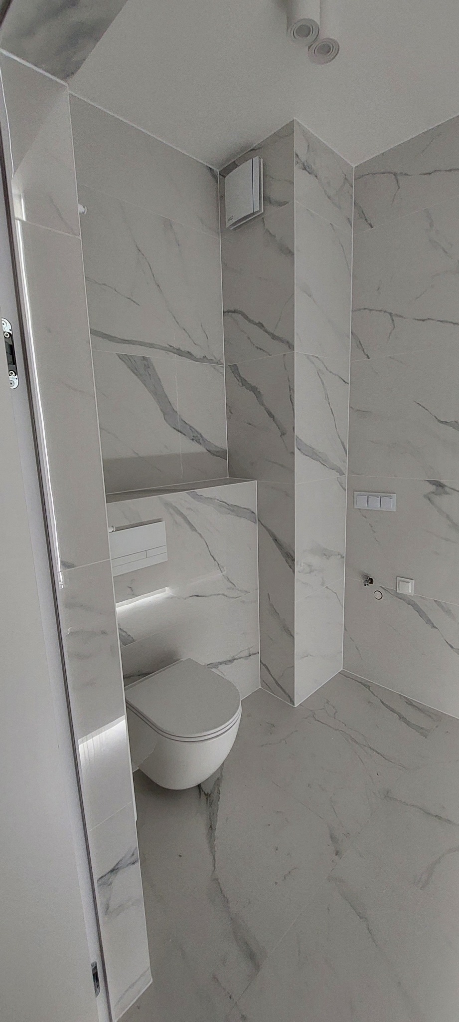 Nowoczesna, minimalistyczna łazienka wykończona białymi płytkami imitującymi marmur, z podwieszaną toaletą i subtelnym oświetleniem LED pod półką.