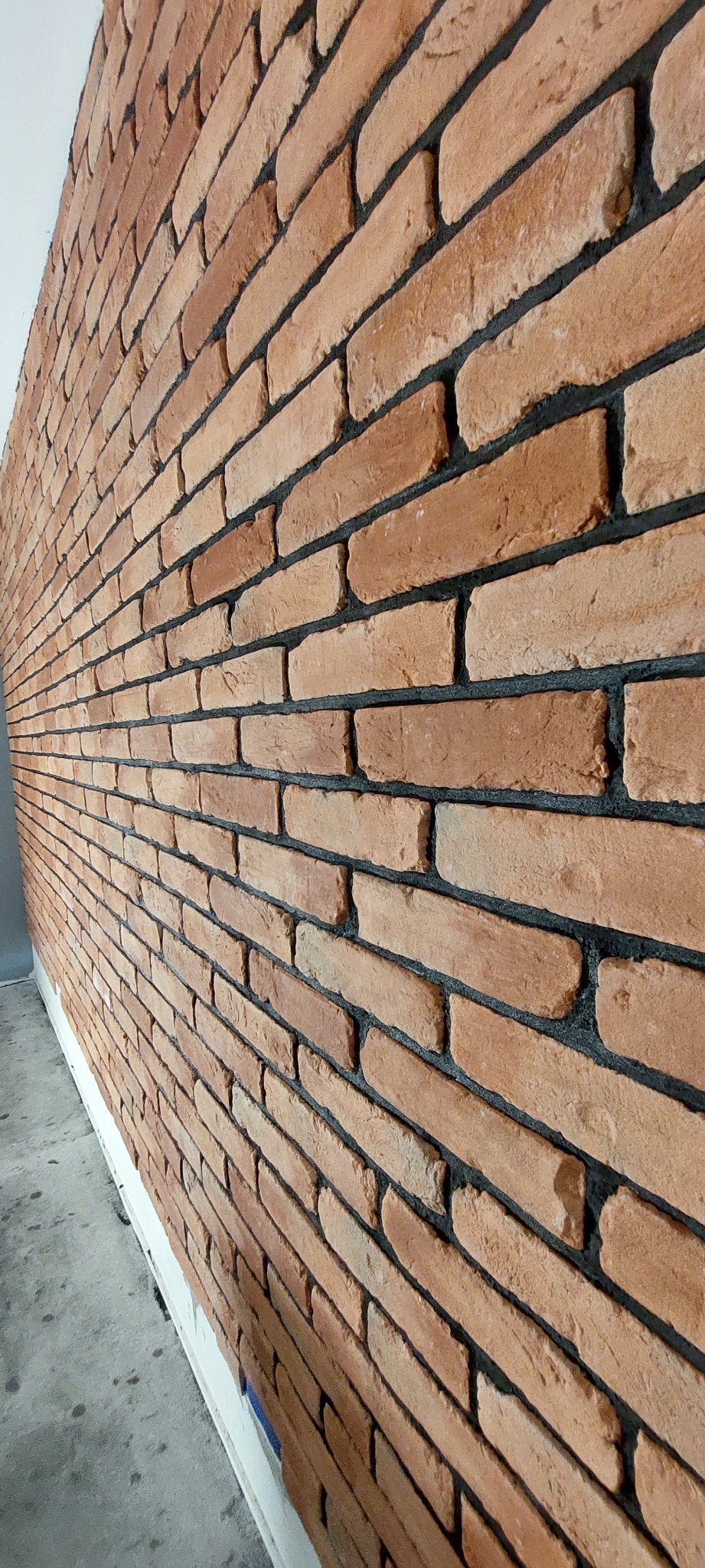 Ściana wyłożona cegłą klinkierową w odcieniach czerwieni i brązu, z ciemną fugą, widoczna z perspektywy bocznej, z fragmentem szarej podłogi i białej listwy przypodłogowej.