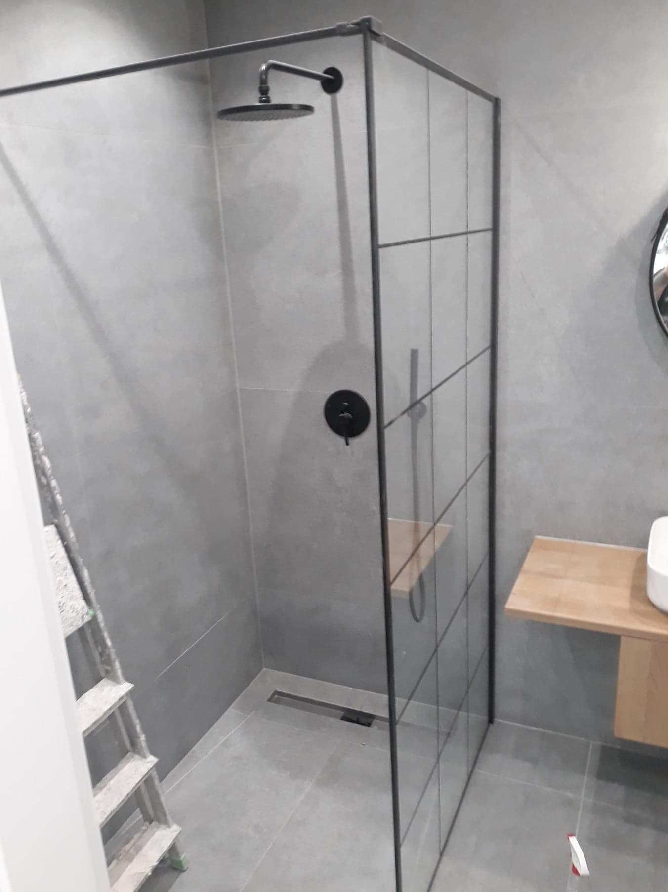Nowoczesna kabina prysznicowa walk-in z czarnymi profilami na tle szarych płytek, widoczna drabina malarska i drewniana półka z umywalką.