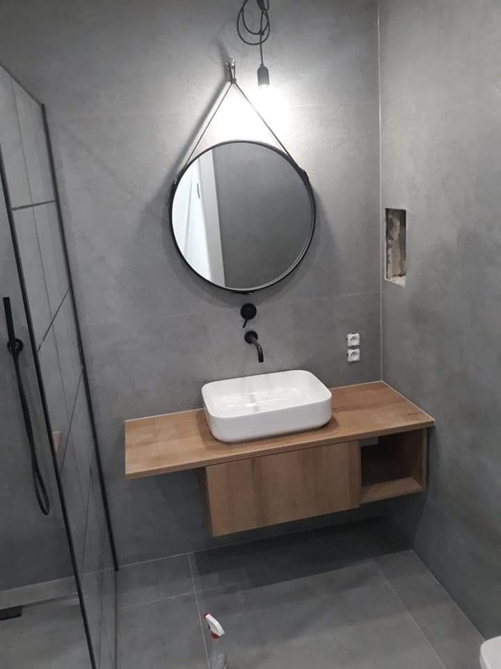 Nowoczesna łazienka z szarą glazurą, okrągłym lustrem na pasku, białą umywalką nablatową na drewnianym blacie i czarną armaturą, widoczny fragment kabiny prysznicowej.