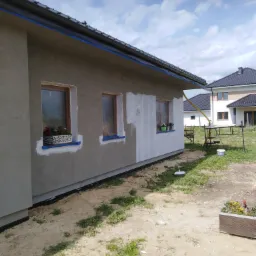 Fragment ściany domu jednorodzinnego w trakcie prac przygotowawczych do malowania, z oknami zabezpieczonymi taśmą malarską i kwiatami w doniczkach na parapetach, częściowo zagruntowany fragment...