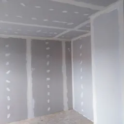 Wnętrze pokoju z zamontowanymi płytami kartonowo-gipsowymi, spoiny między płytami zaszpachlowane i zaszlifowane, widoczne ślady po wkrętach na ścianach i suficie, surowa podłoga z płyt OSB.