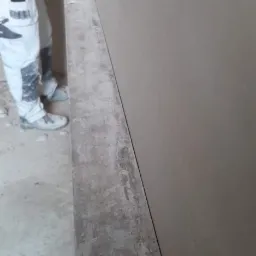 Świeżo nałożony tynk maszynowy na ścianie, z widocznym pracownikiem używającym poziomicy laserowej, w tle robocze spodnie z kieszeniami.