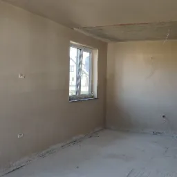 Wnętrze surowego pokoju po nałożeniu tynku, widoczne okna, surowy betonowy strop oraz przygotowane materiały budowlane na podłodze.