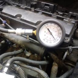 DB Service Dariusz Bryl - Manometr podłączony do silnika samochodowego podczas diagnostyki w warsztacie, widoczne przewody i elementy silnika w zbliżeniu.