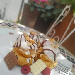 Trzy ptysie z białym lukrem, posypane płatkami migdałów i polane czekoladą, udekorowane kostkami czekolady (mlecznej i białej) oraz malinami, na białym talerzu z geometrycznym wzorem na mozaikowym...