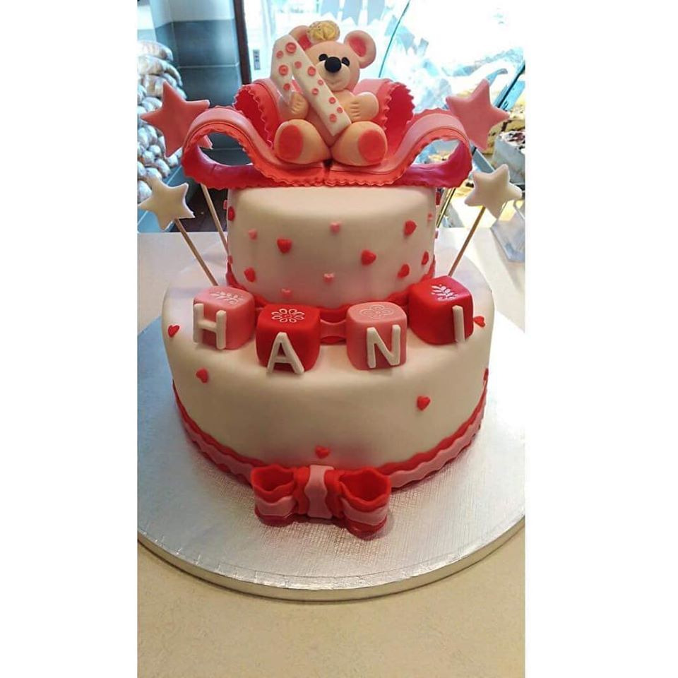 Dwupiętrowy tort z białym lukrem, ozdobiony czerwonymi serduszkami, kokardą i kostkami z literami układającymi się w imię 'HAN', z figurką misia na szczycie i gwiazdkami na patyczkach.