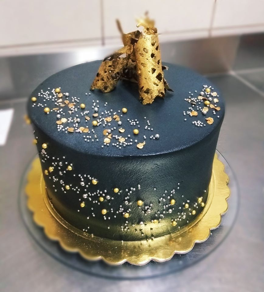 Elegancki tort o ciemnym, grafitowym kremie, ozdobiony złotymi i srebrnymi kuleczkami oraz dwoma złotymi, ażurowymi elementami na wierzchu, umieszczony na złotej podkładce.