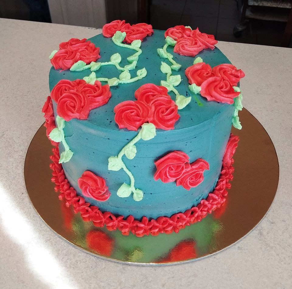 Okrągły tort z turkusowym kremem, zdobiony czerwonymi różami i zielonymi pnączami z kremu, umieszczony na złotej podkładce.