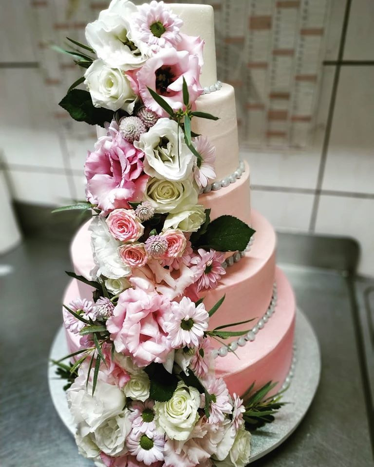 Czteropiętrowy tort weselny w odcieniach różu i bieli, udekorowany kaskadą świeżych kwiatów, w tym róż, eustom i goździków, oraz sznurem srebrnych perełek.
