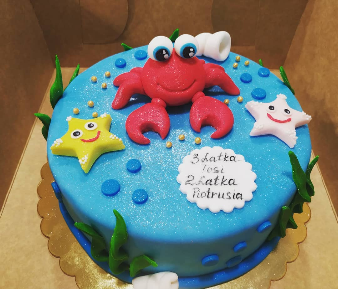 Tort urodzinowy z motywem morskim: niebieski lukier, krab i gwiazdy morskie z lukru, napis '3 latka Tosi, 2 latka Piotrusia' na białej dekoracji.