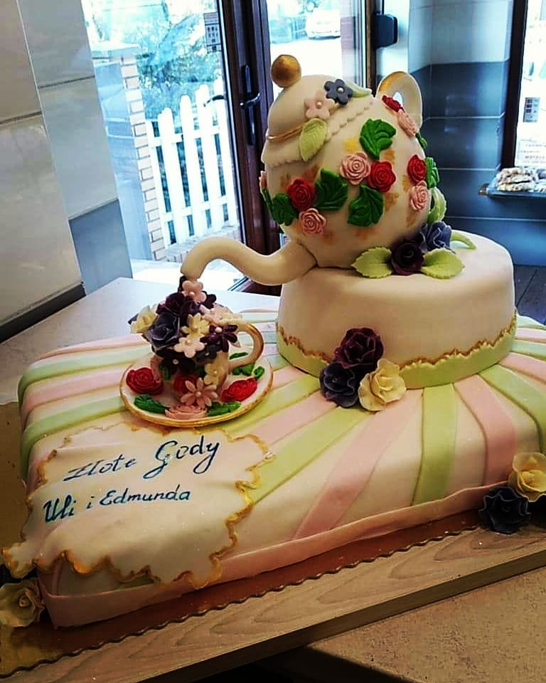 Wielopoziomowy tort w kształcie imbryka i filiżanki, zdobiony kwiatami i napisem 'Złote Gody Uli i Edmunda', na tle okna.