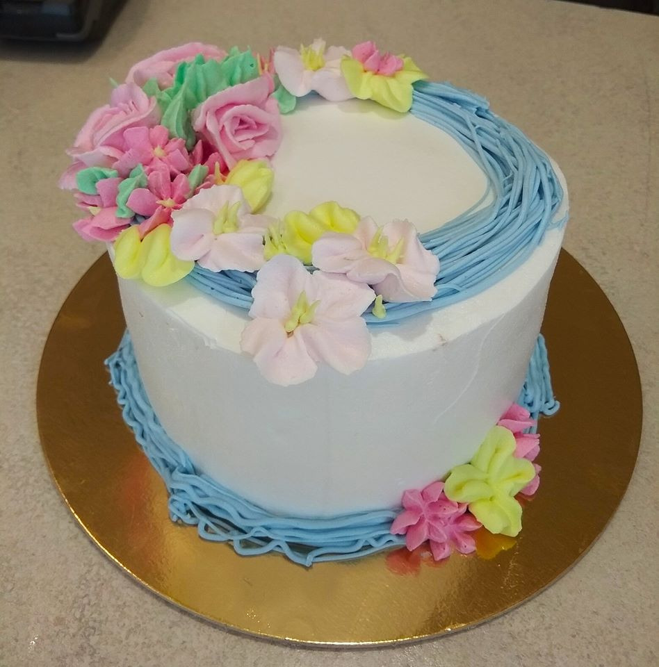 Tort z białym kremem, dekorowany niebieskim kremem w kształcie gniazda i różowymi, żółtymi oraz zielonymi kwiatami z kremu, umieszczony na złotej podkładce.
