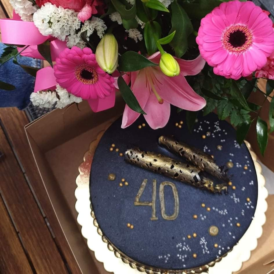 Tort na 40. urodziny z ciemnym lukrem, złotymi ozdobami w kształcie cygar i napisem '40', udekorowany bukietem różowych kwiatów i lilii.