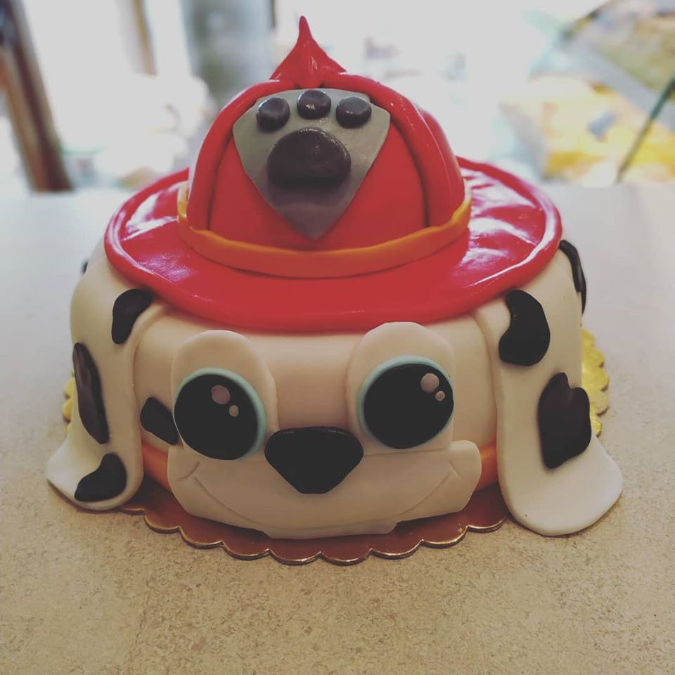 Tort urodzinowy w kształcie psa z bajki, z czerwonym hełmem strażackim na głowie i dużymi, okrągłymi oczami, na złotym podkładzie.