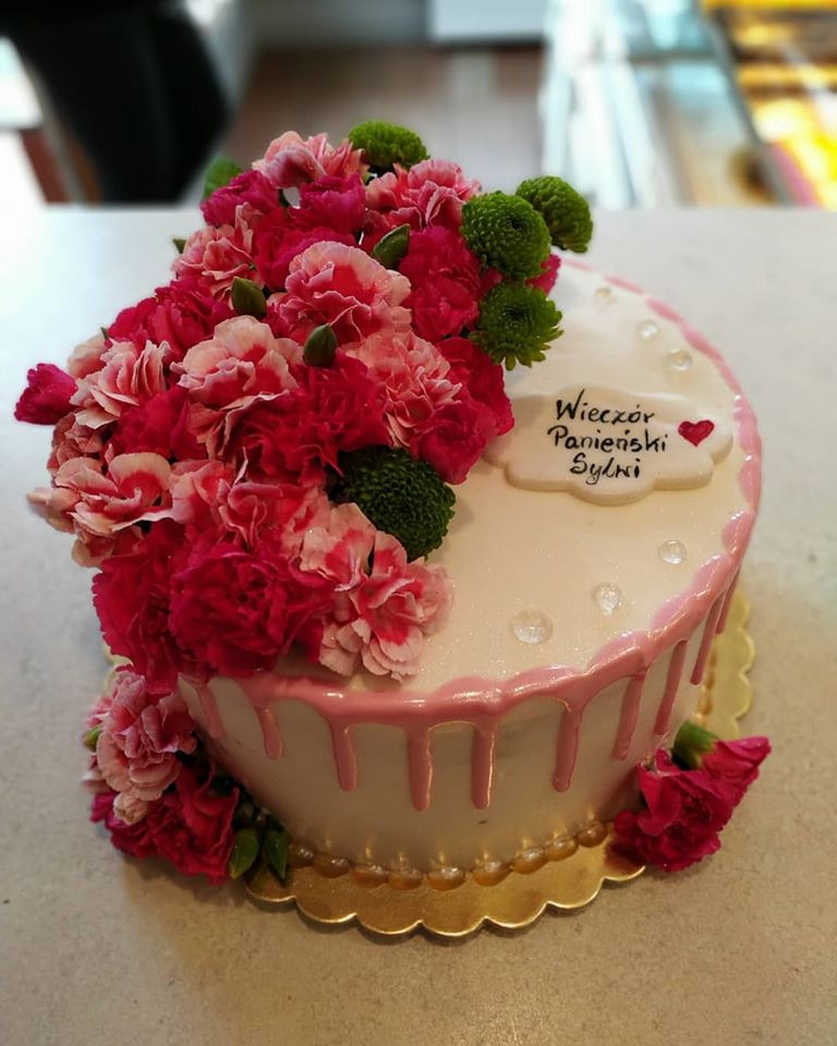 Tort z białym lukrem, różowymi polewami i dekoracją z różowych i zielonych kwiatów, dedykacja na wieczór panieński.